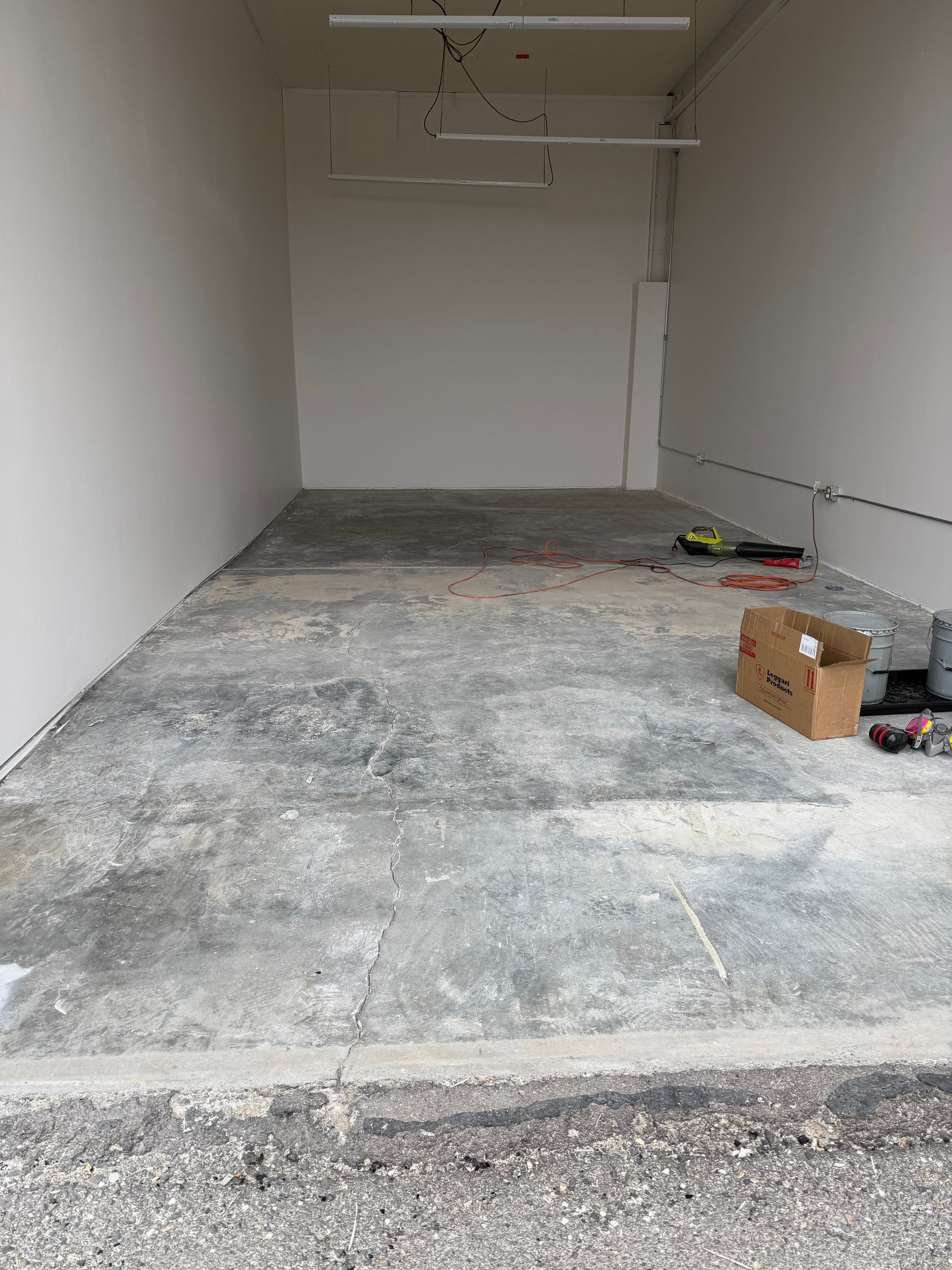 An unfinished indoor space with bare white walls, a concrete floor, and overhead lighting, showing signs of construction.