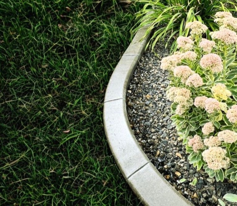 A curved stone garden border separates green grass from a flower bed filled with dark gravel and blooming pale flowers.