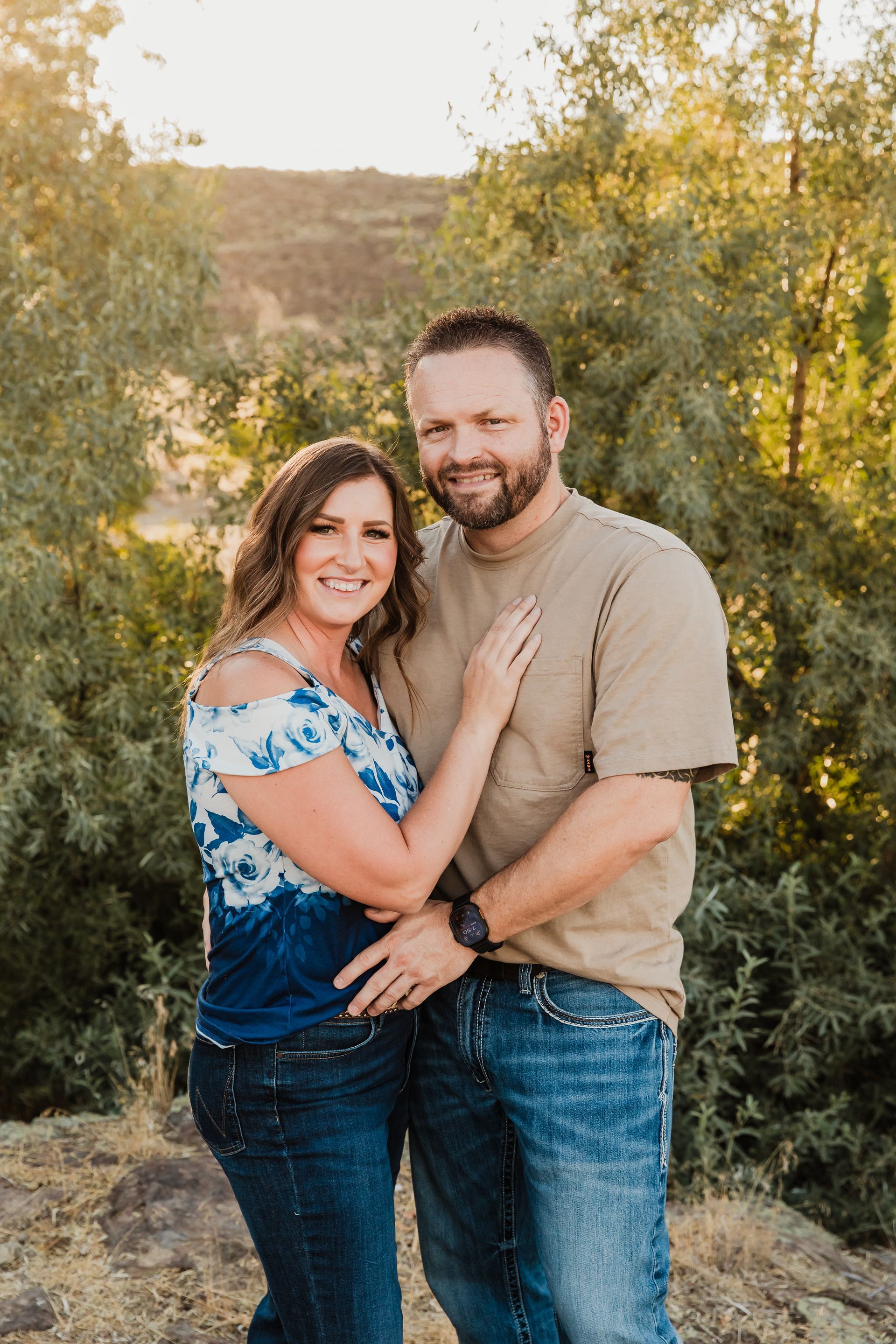 A smiling couple stands in an outdoor, wooded area with golden sunlight, embracing while looking toward the camera.