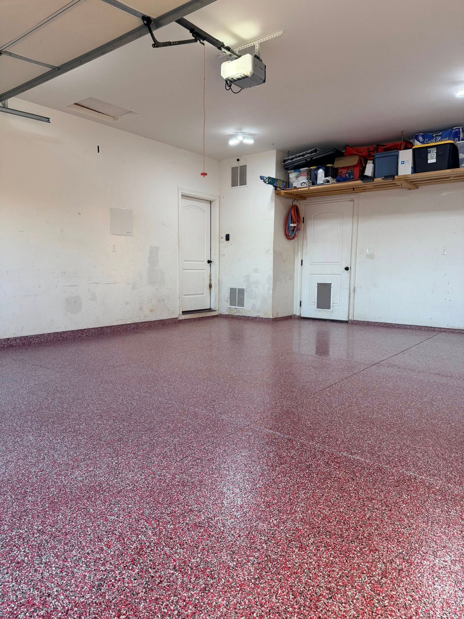 A garage with a shiny, red and white speckled epoxy floor, white walls, two white doors, and a storage shelf.