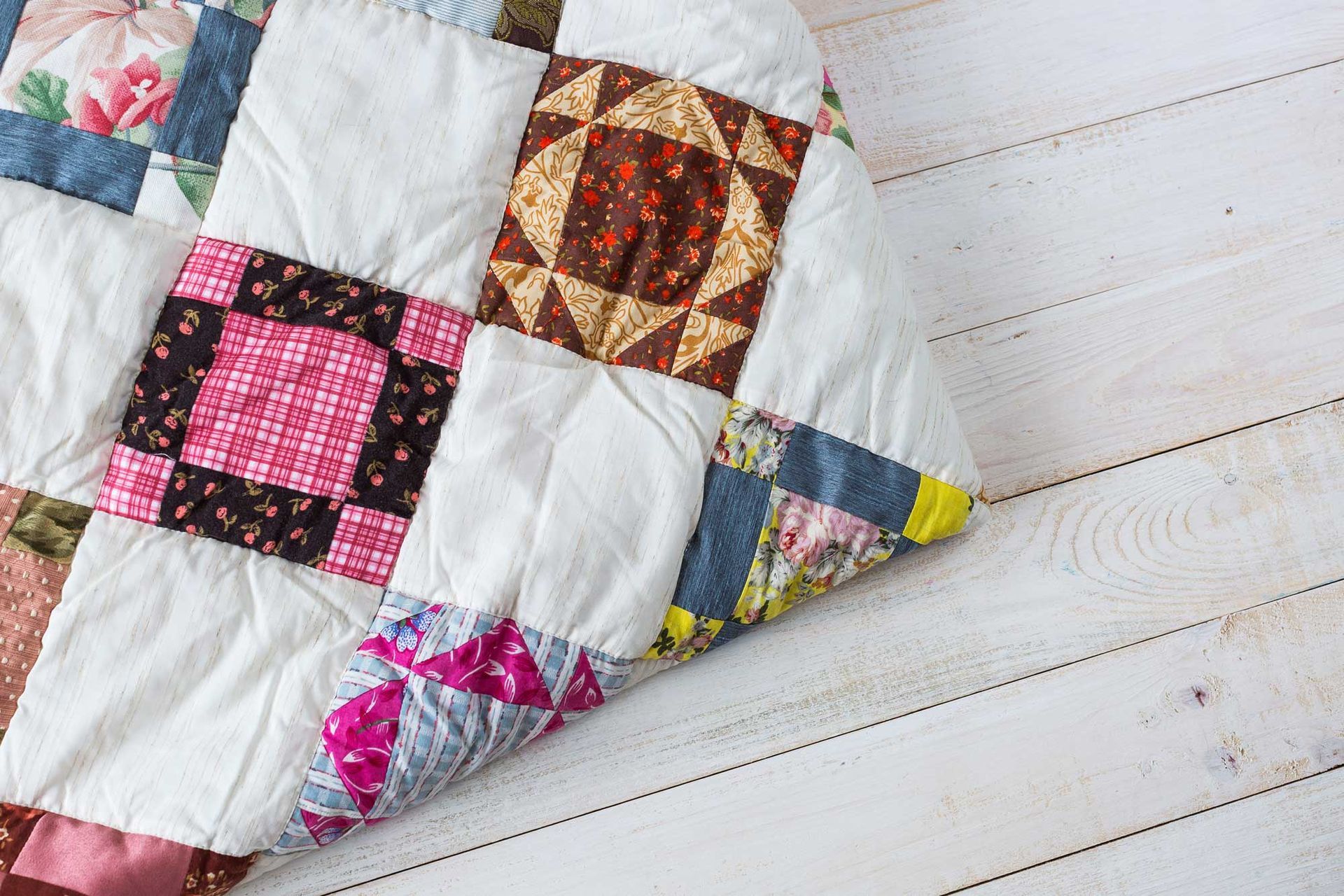 A folded patchwork quilt with colorful patterned fabric squares on a light wooden floor.