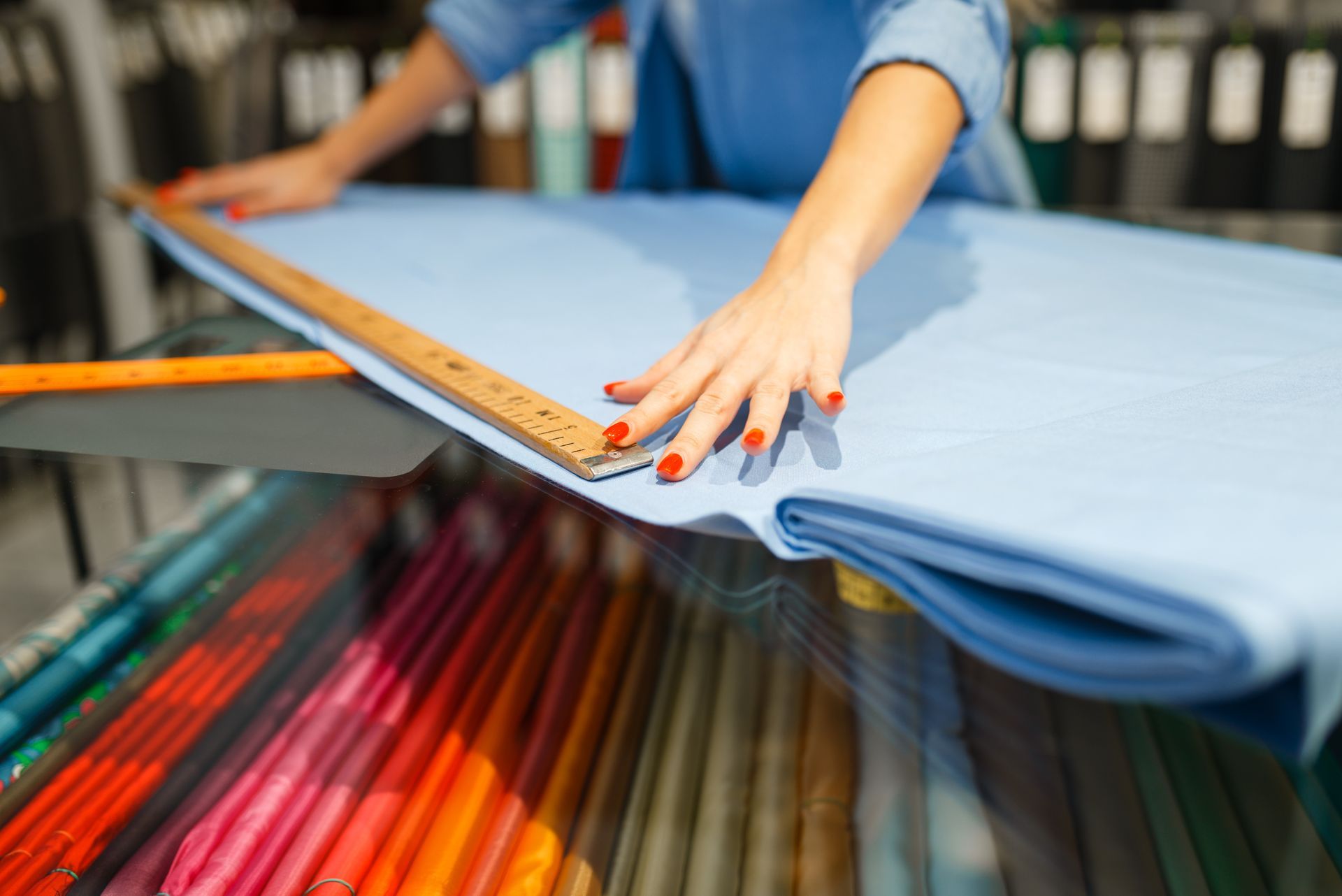 Hands measuring folded blue fabric with a ruler on a table, surrounded by colorful fabric rolls.