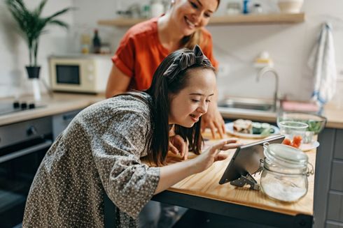 A woman and a girl are looking at a tablet in a kitchen.