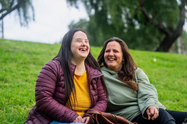 Two women are sitting on the grass in a park laughing.
