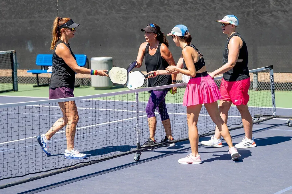Four women shaking hands over a pickleball net after a game on an outdoor court. 