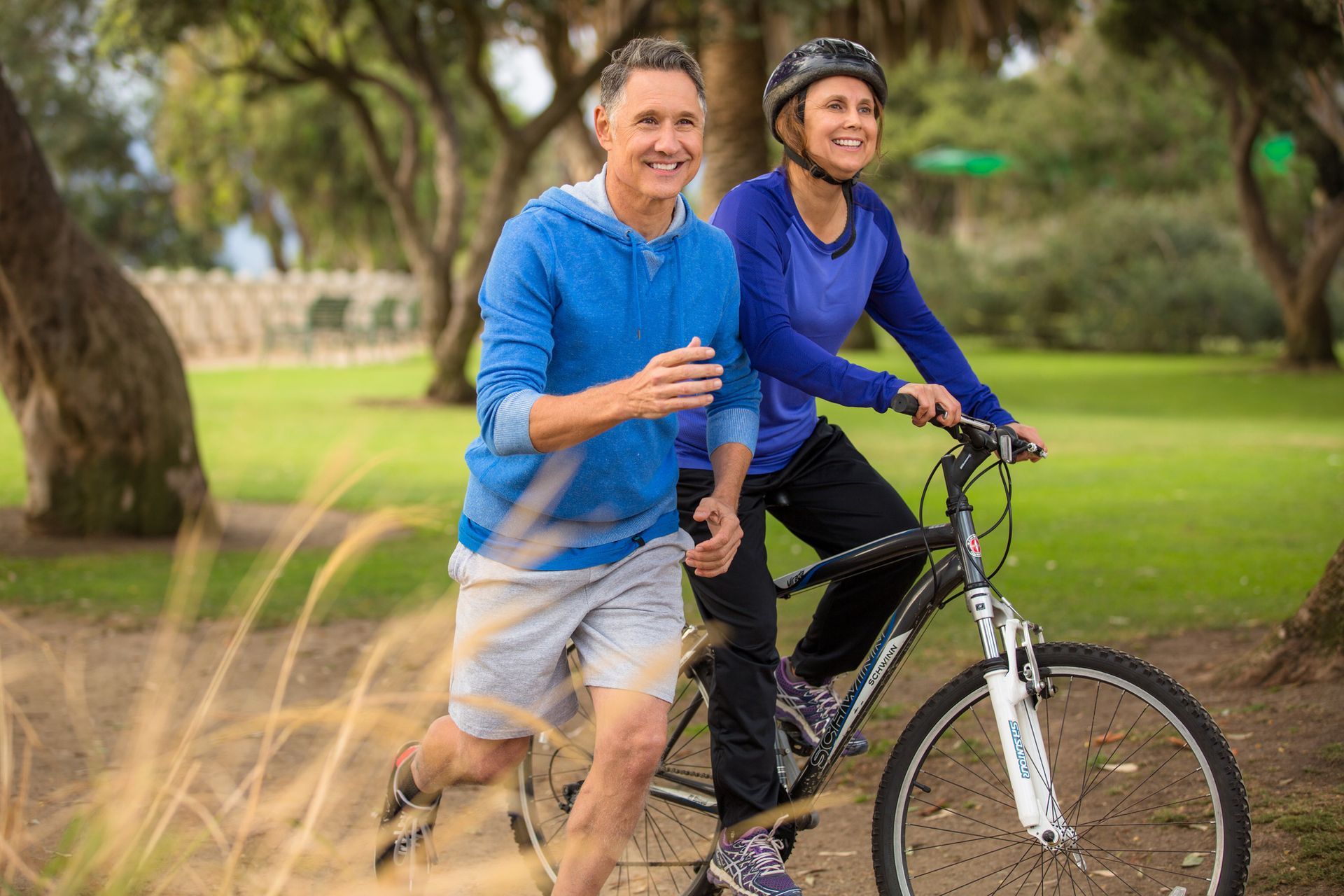 Man jogging next to a woman riding a bicycle in a park. They are smiling, with trees and greenery in the background.