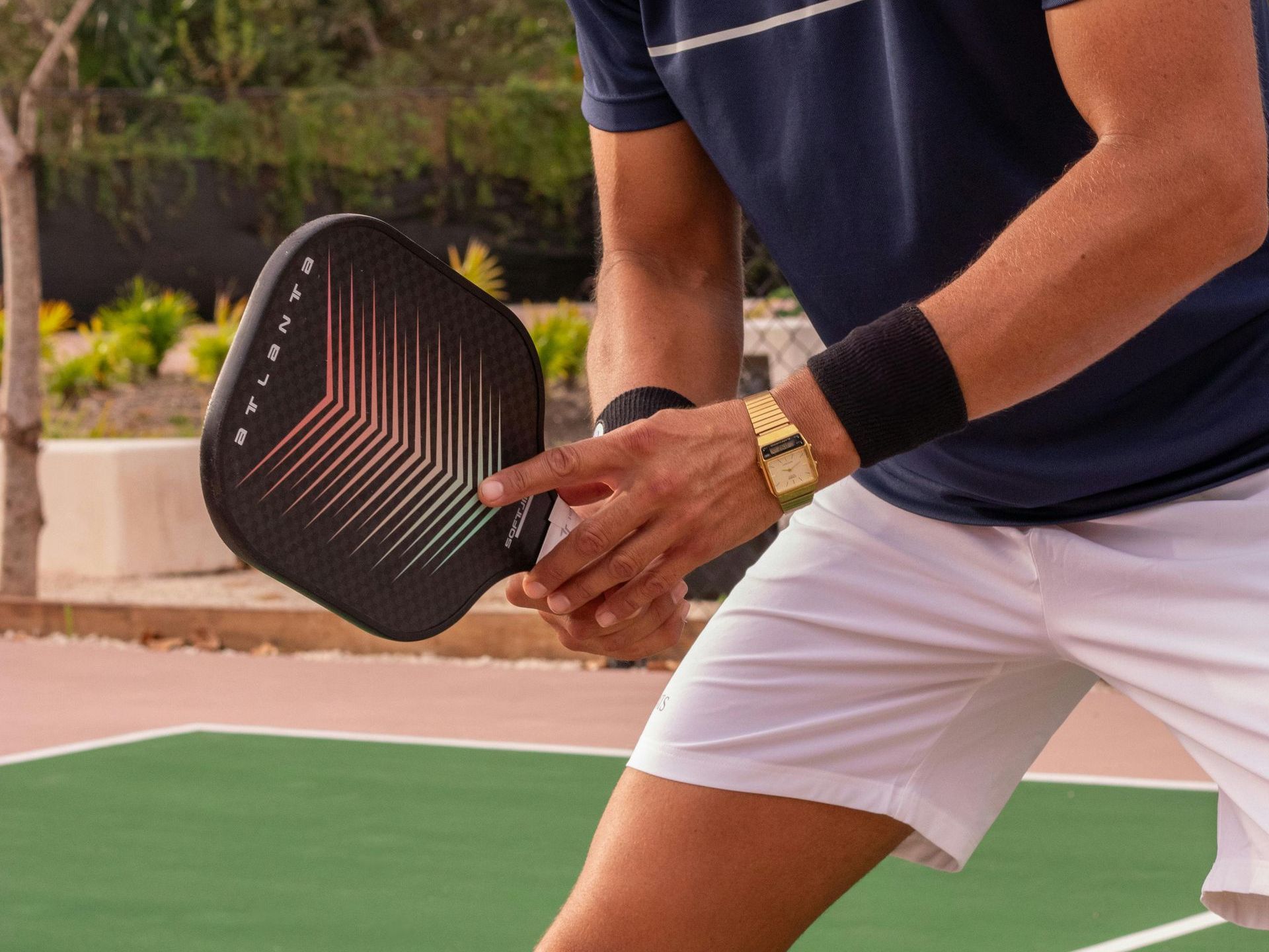 A person playing pickleball, holding a paddle with a red and green design, wearing a gold watch and wristband.