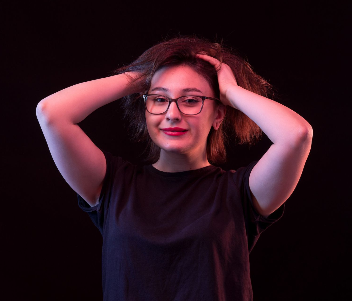 Una mujer con gafas sonríe, con las manos en el cabello, iluminada por una luz rosa y azul sobre un fondo negro.