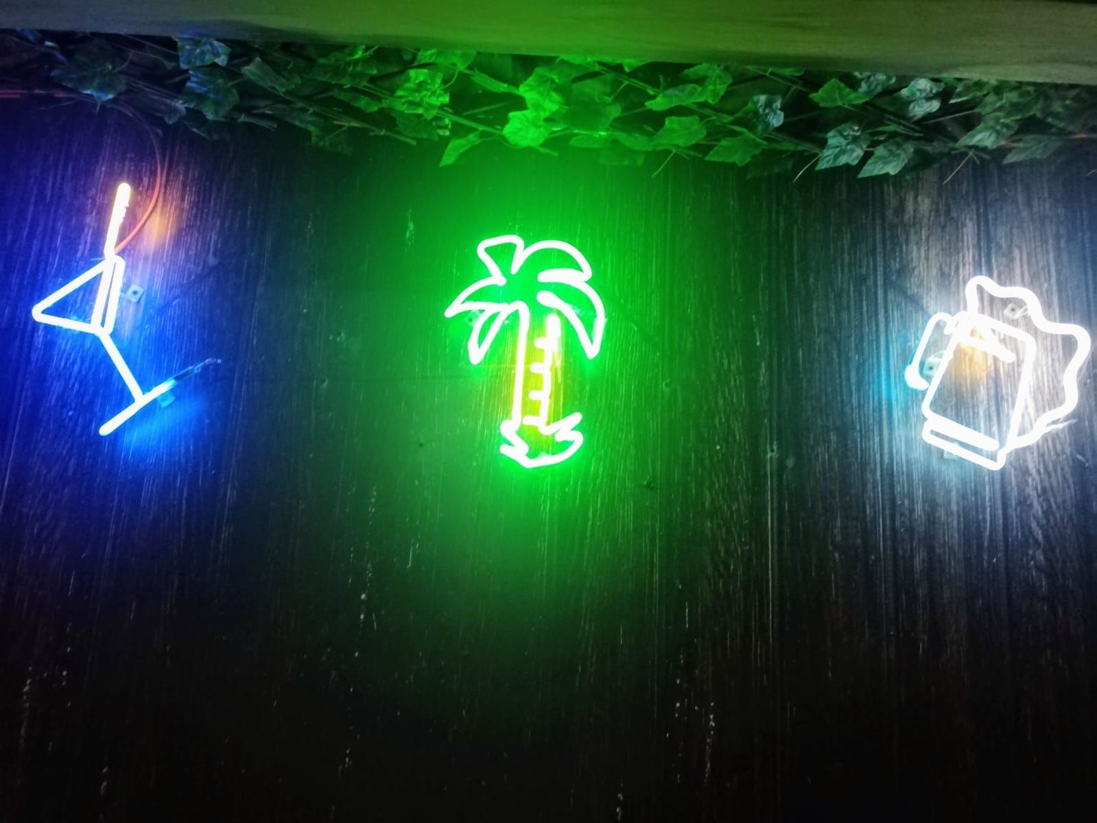 Una mujer con gafas sonríe, con las manos en el cabello, iluminada por una luz rosa y azul sobre un fondo negro.