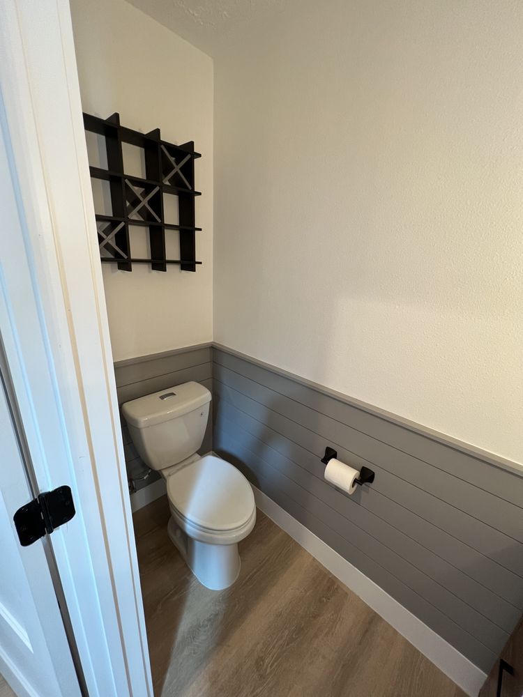 Small bathroom with a white toilet, gray wainscoting, wall shelf, and toilet paper holder.