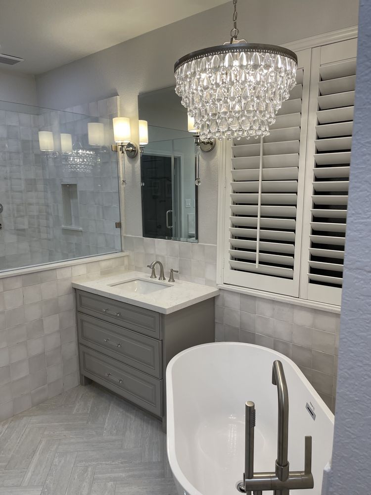 Bright white bathroom with crystal chandelier, gray vanity, marble tiles, and freestanding tub.
