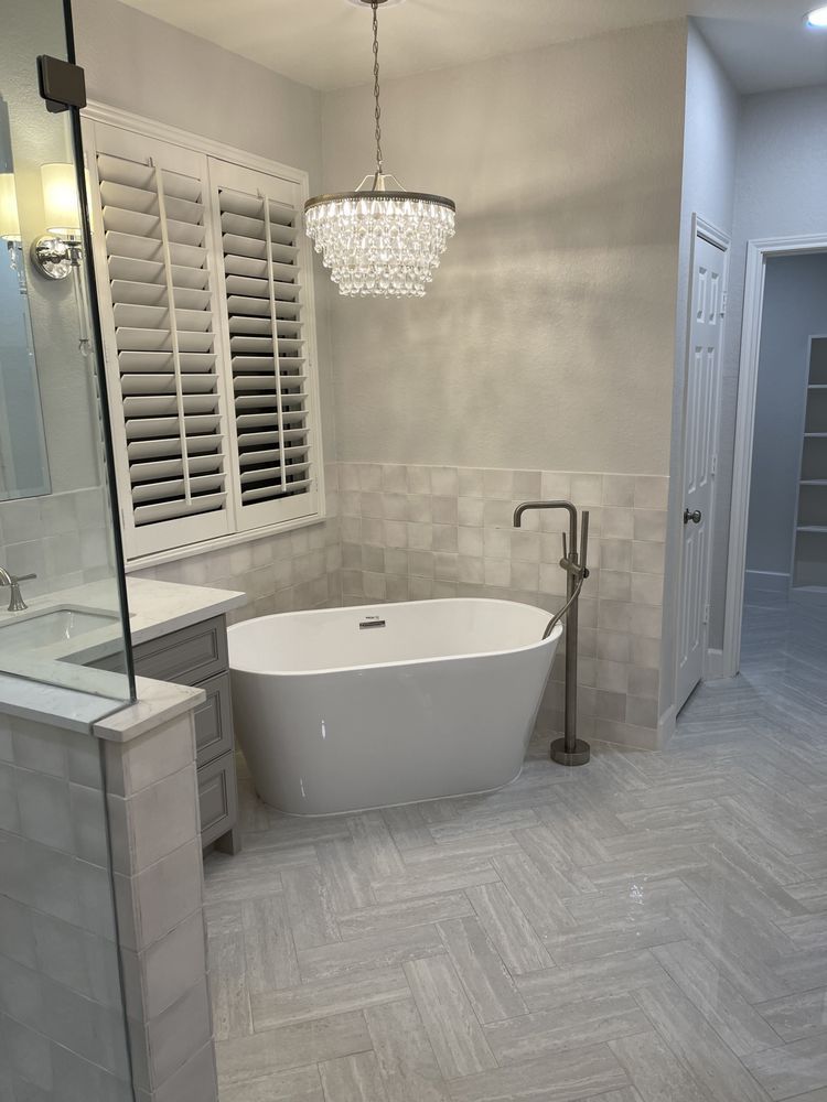 Bright modern bathroom with freestanding tub, chandelier, herringbone floor, and glass shower panel