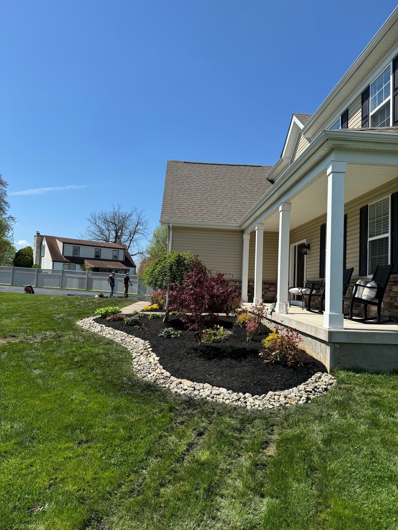 House with porch, flower bed with dark mulch, and rocks edging. Green lawn, blue sky.