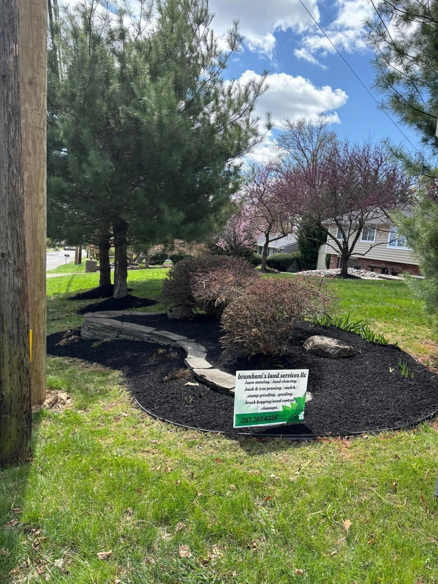 Landscaped yard with dark mulch, shrubs, and a path. Sign on grass, trees, and cloudy blue sky.