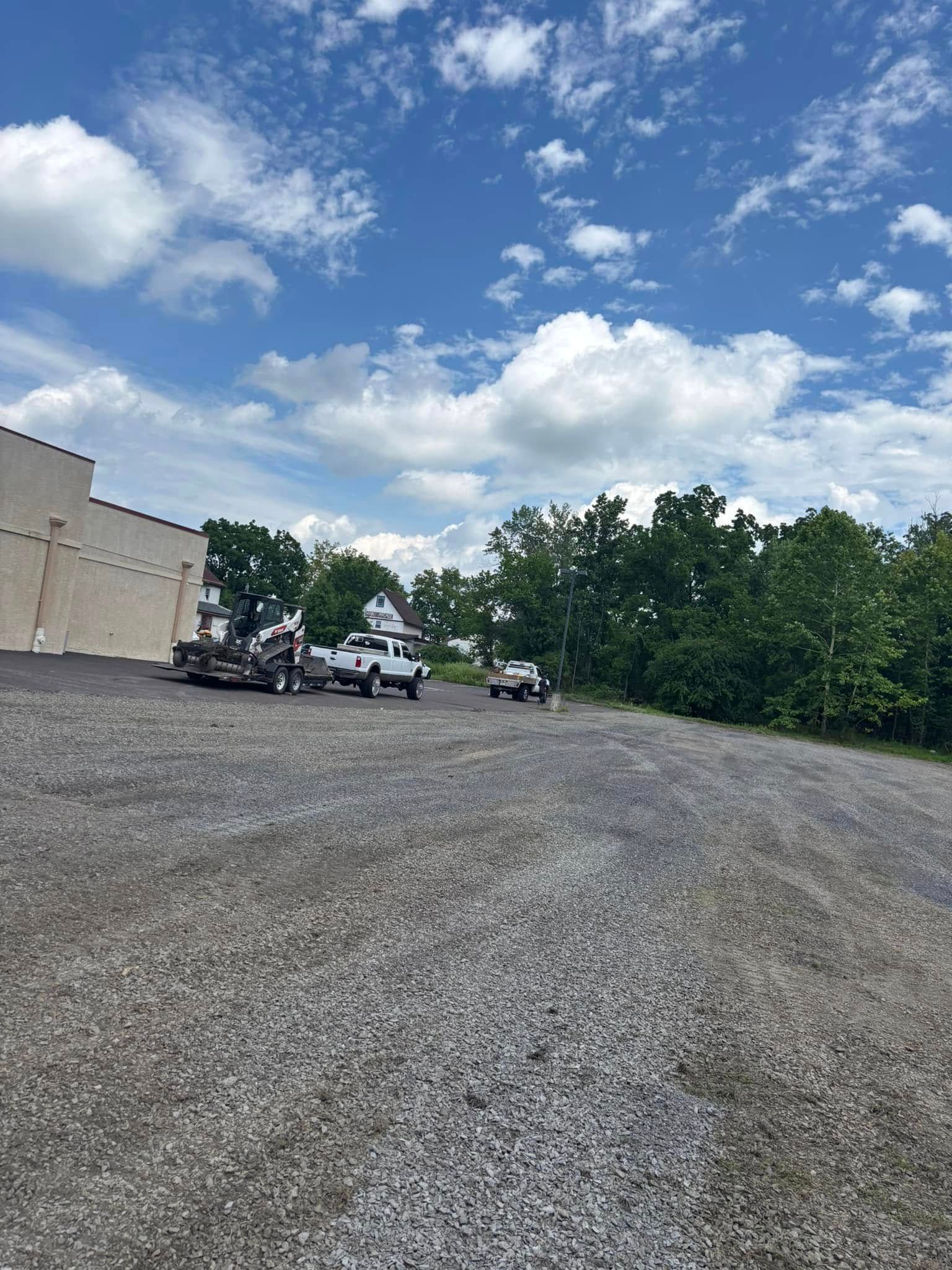 Gravel lot with two white trucks, one towing a trailer, under a blue sky with fluffy clouds.