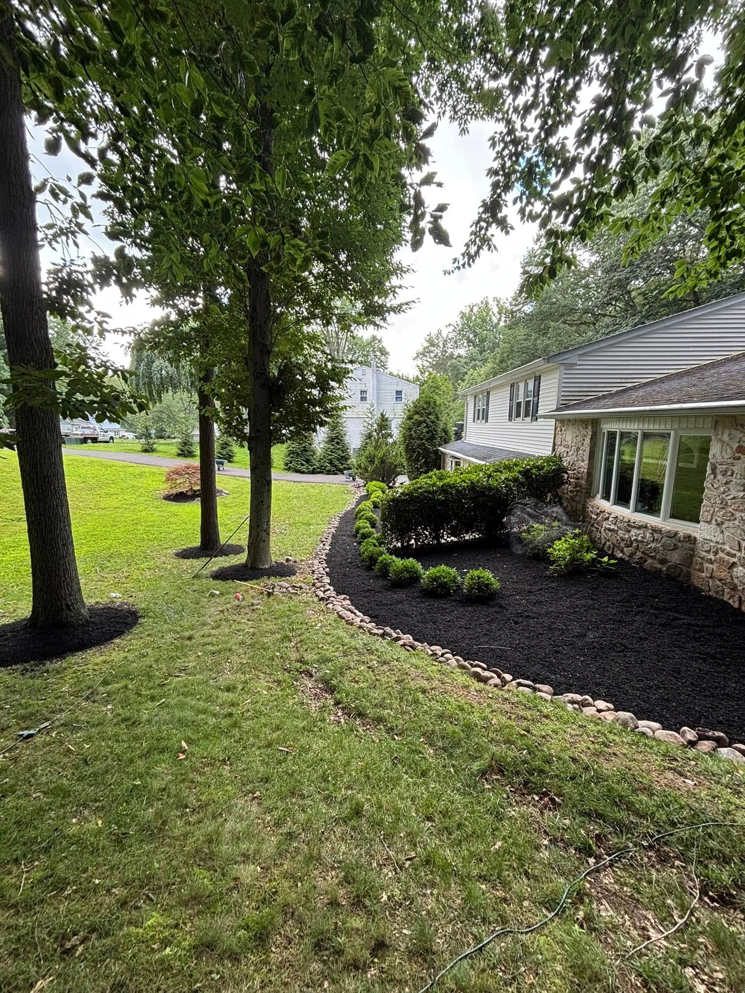 Lawn with trees, mulch bed, and a house with a stone facade under a cloudy sky.