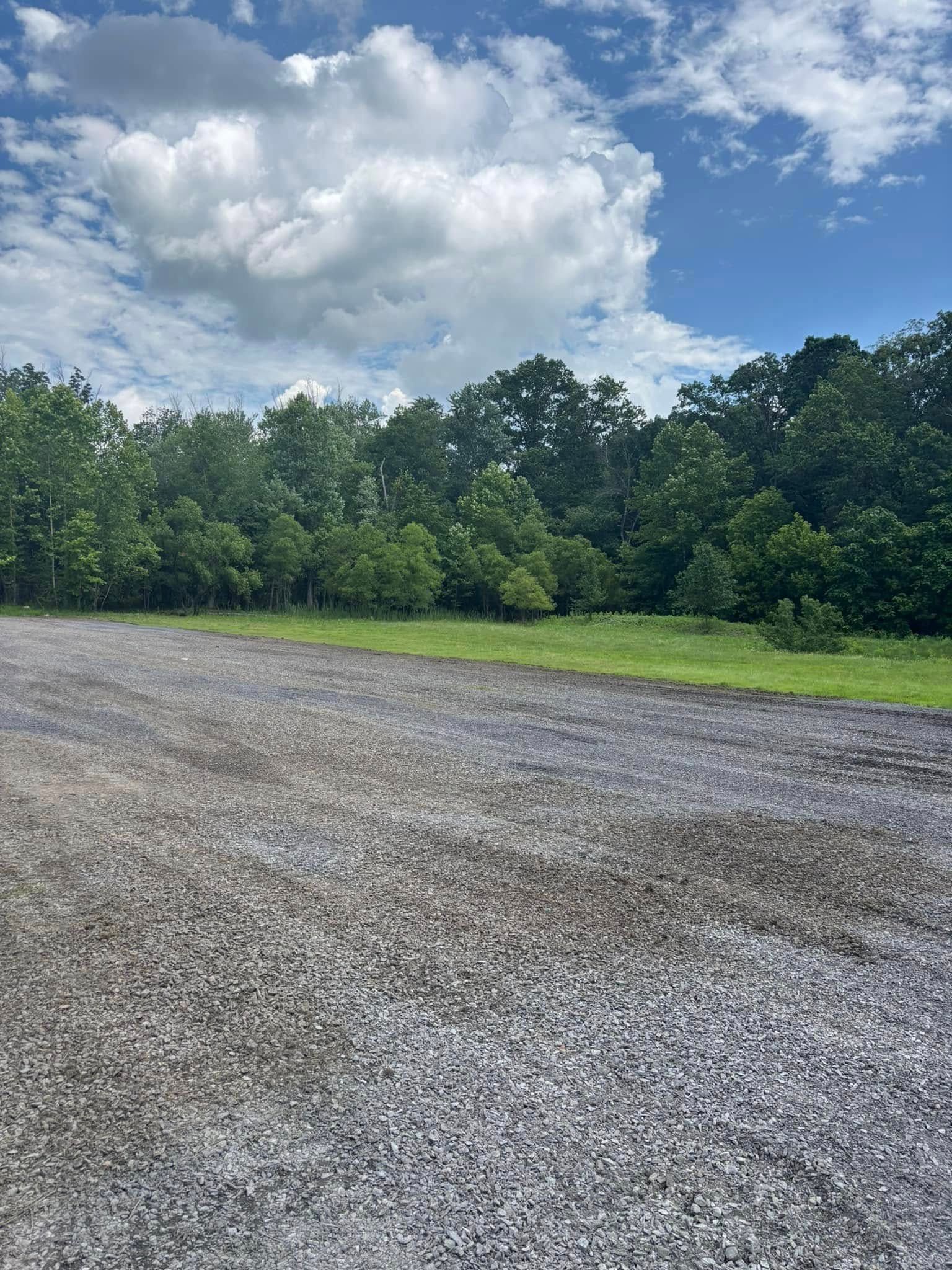 Gravel area with grass and trees under a cloudy blue sky.