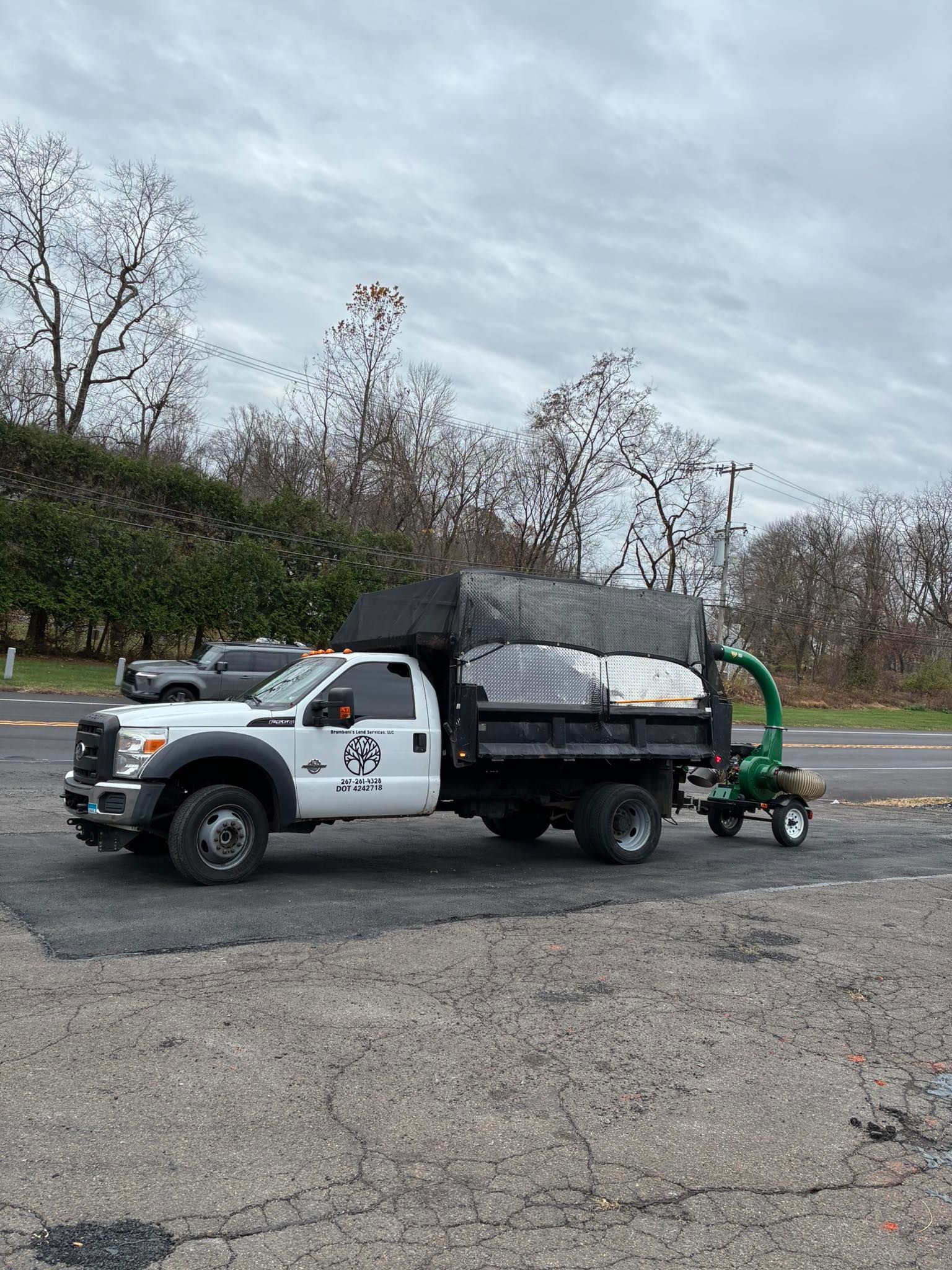 White work truck with a black dump bed, pulling a green wood chipper on a road. Cloudy sky.