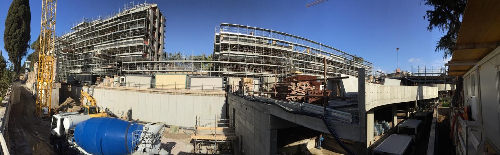 Veduta panoramica di un cantiere edile con strutture in cemento, macchinari pesanti e un'autobetoniera blu.