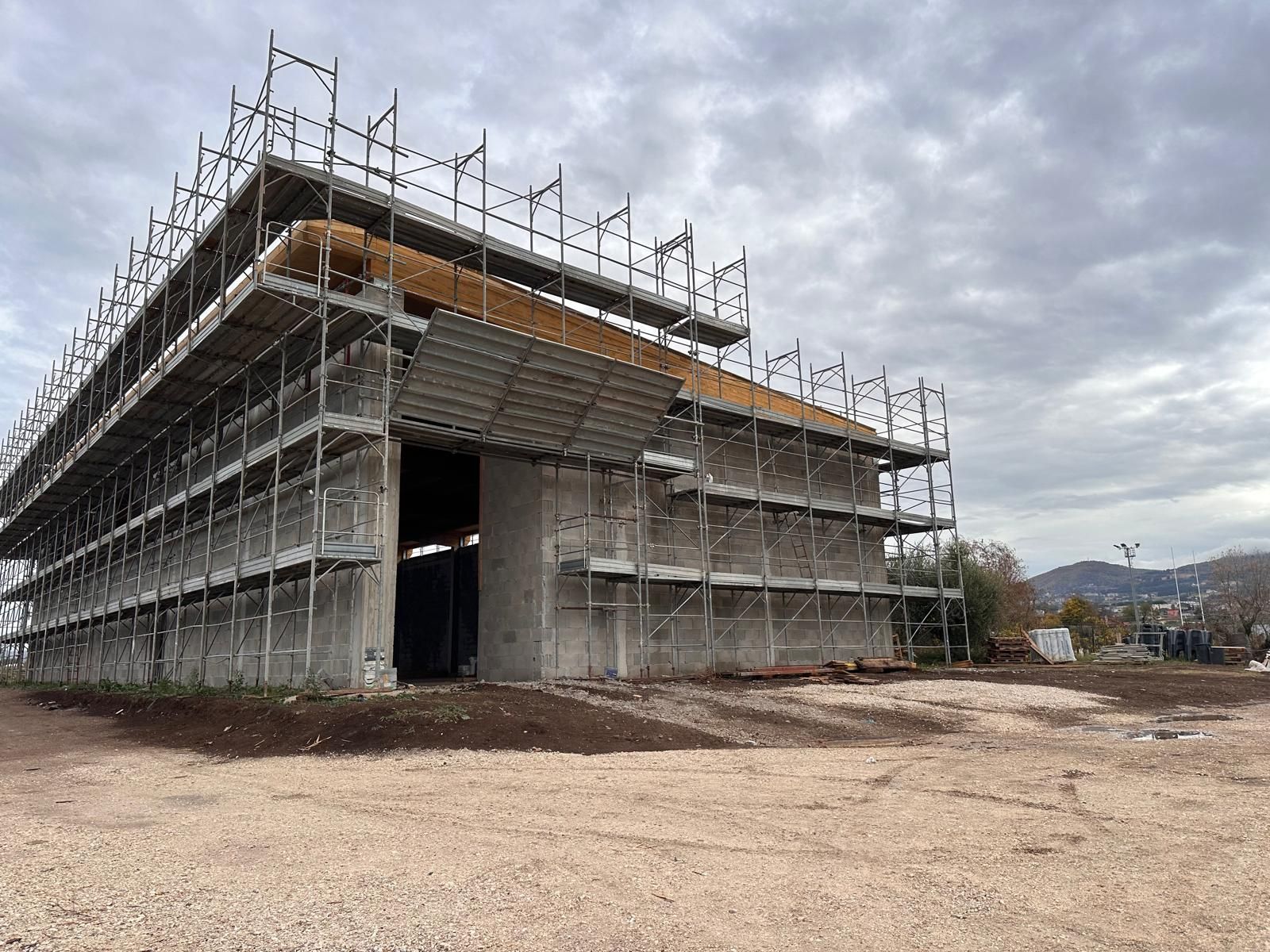 Un edificio in cemento parzialmente costruito, circondato da impalcature metalliche, su un terreno ghiaioso sotto un cielo nuvoloso.