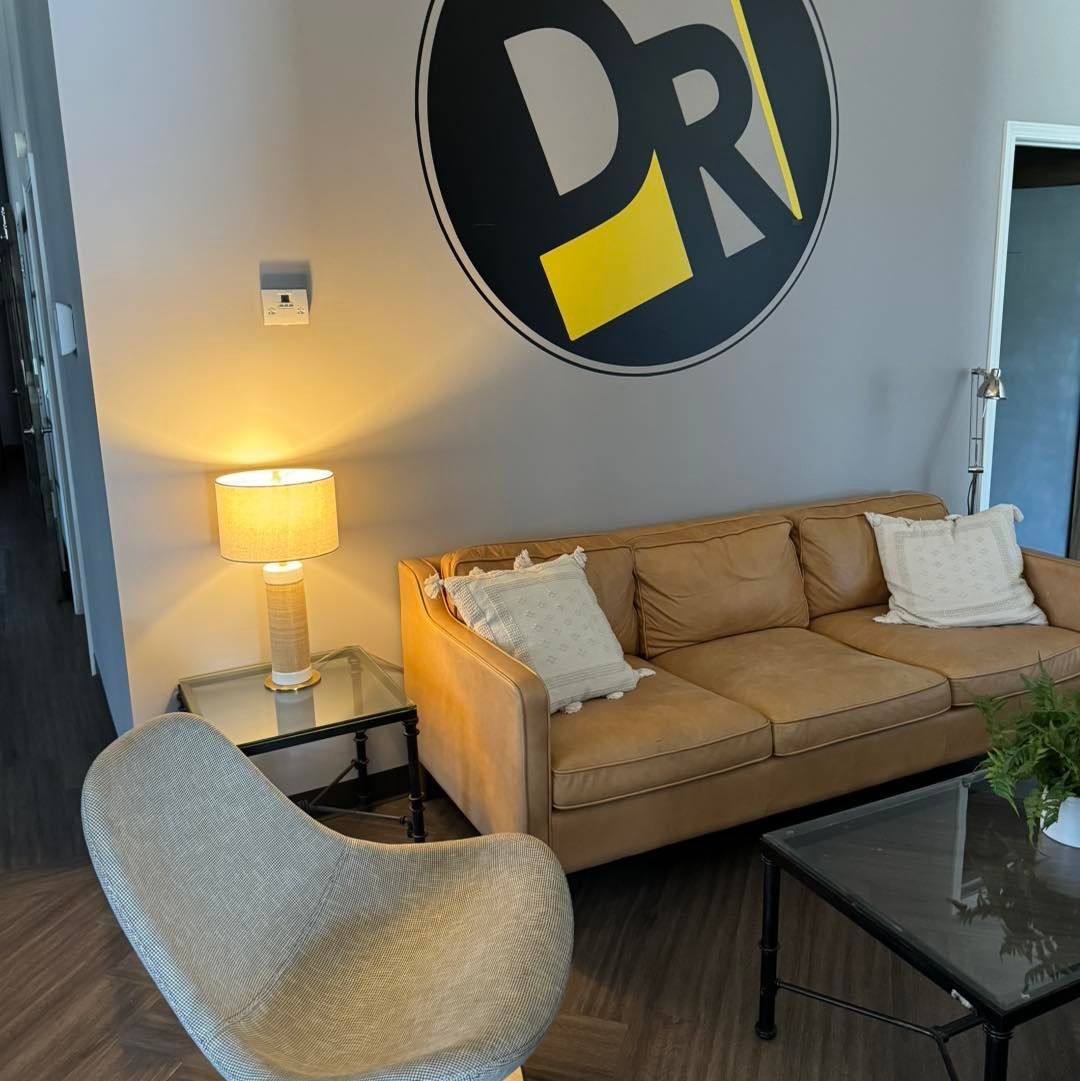 A modern seating area with a tan leather couch, a round side table with a lit lamp, and a chair in a waiting room.