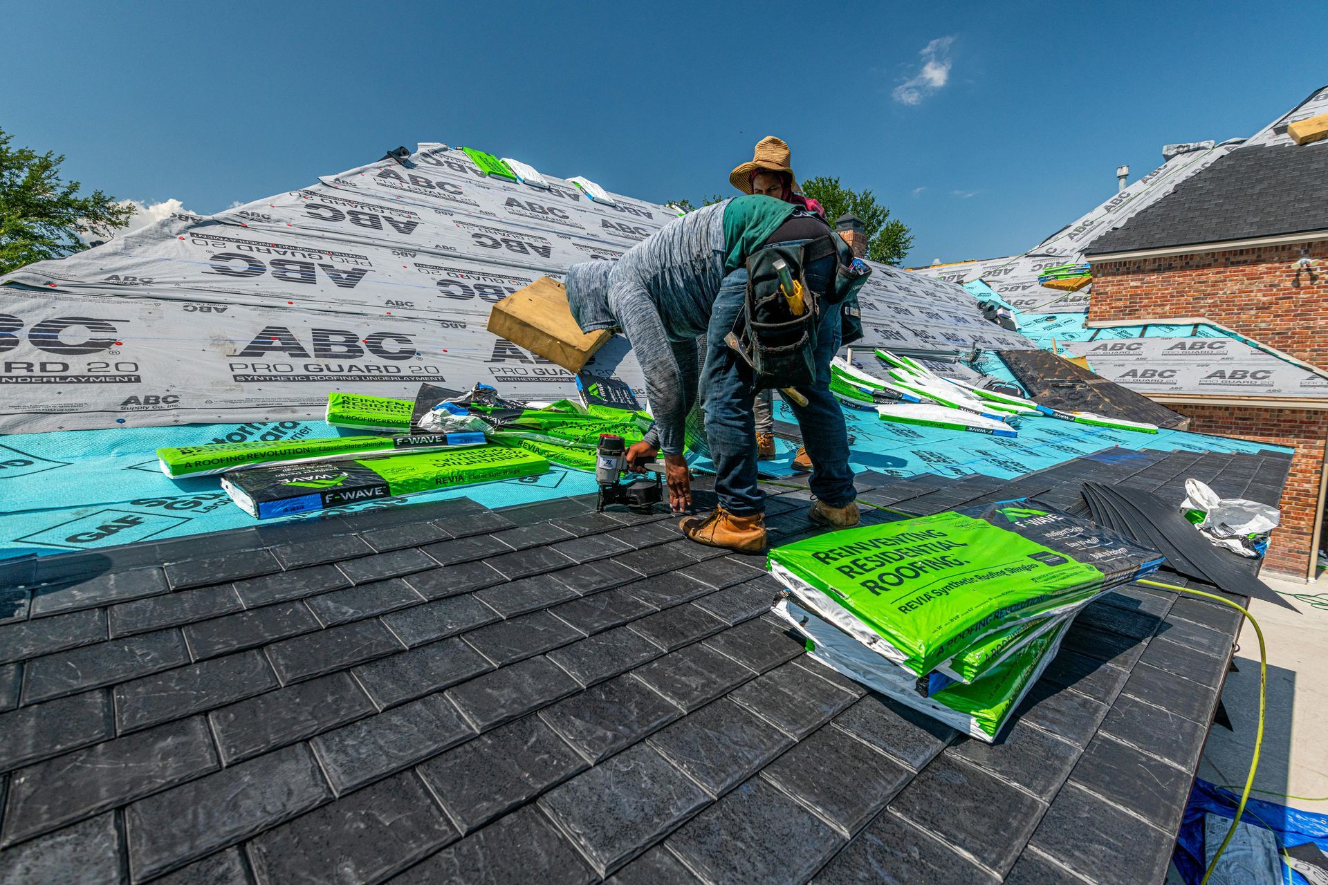 A worker in a tan hat and blue jeans installs black shingles on a house roof under a bright blue sky.