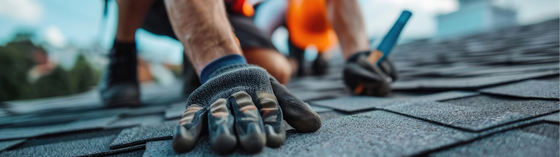 Workers in work gloves and orange vests installing dark asphalt shingles on a roof.