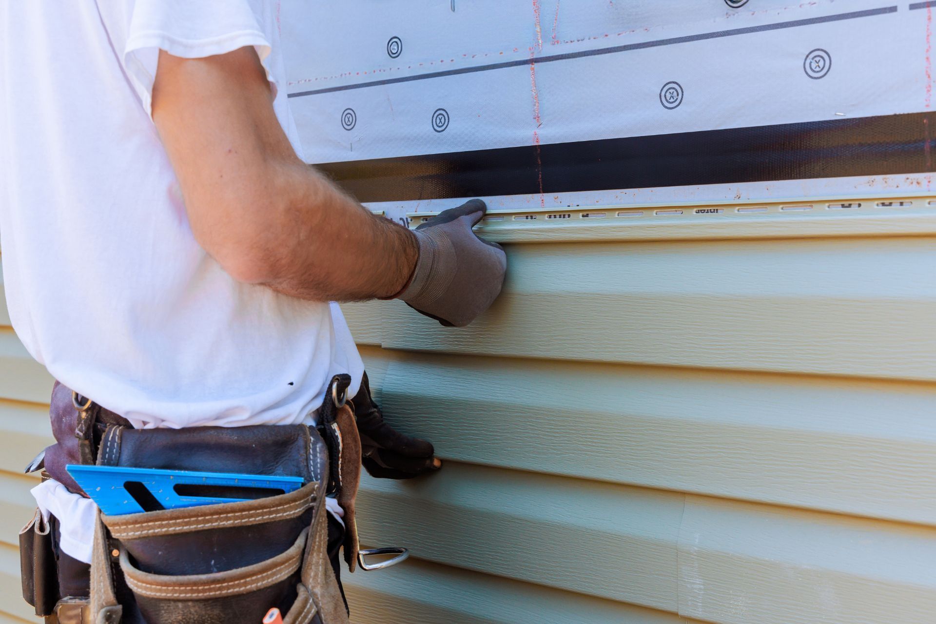 A worker in a white shirt and tool belt installs light green siding on a house wall covered with protective barrier film.