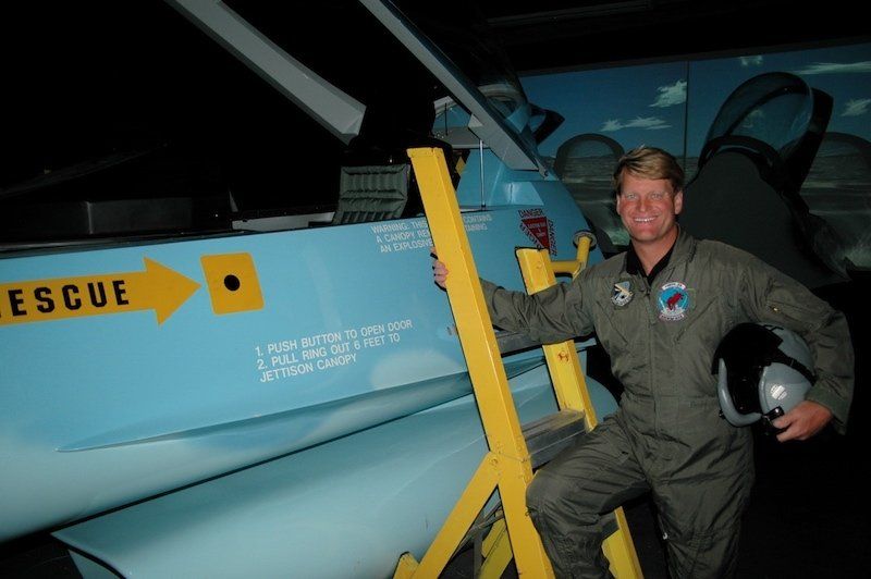 Flightdeck customer Howie Klausner standing at yellow ladder next to F-16 flight simuator