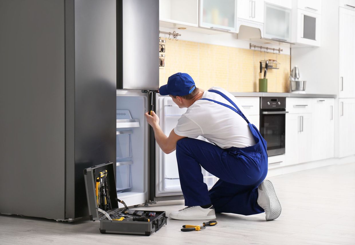 Repair technician in blue overalls fixing a refrigerator in a white kitchen
