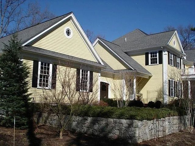 Yellow suburban house with dark gray roof and stone retaining wall on a sunny day