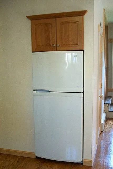 White refrigerator with wood cabinets above in a narrow hallway.