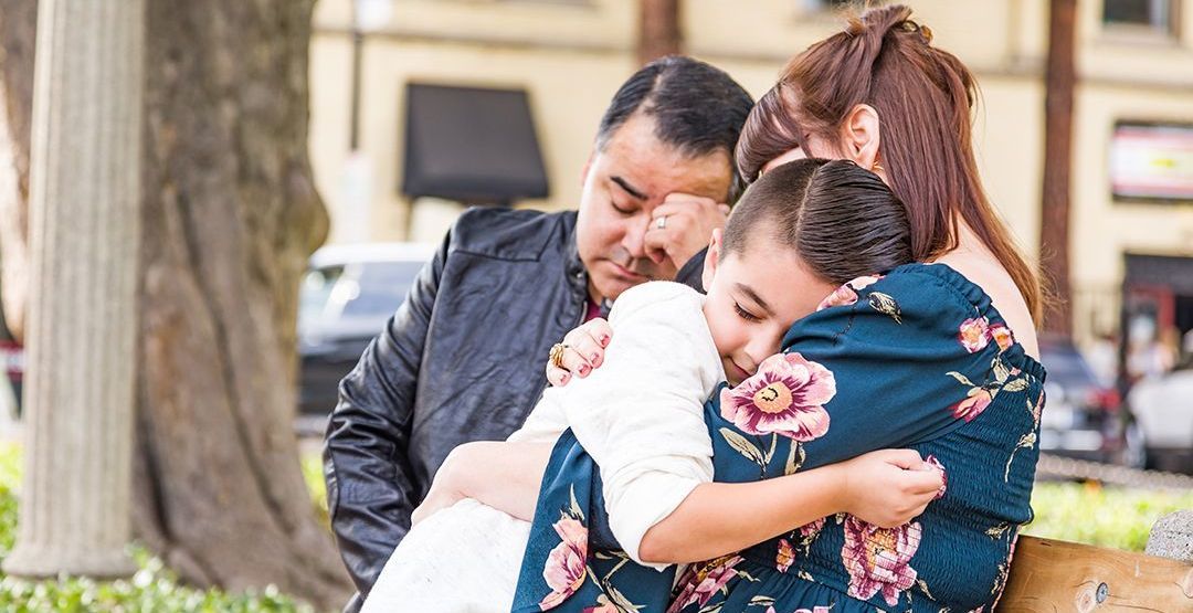 A family is hugging each other while sitting on a bench in a park.