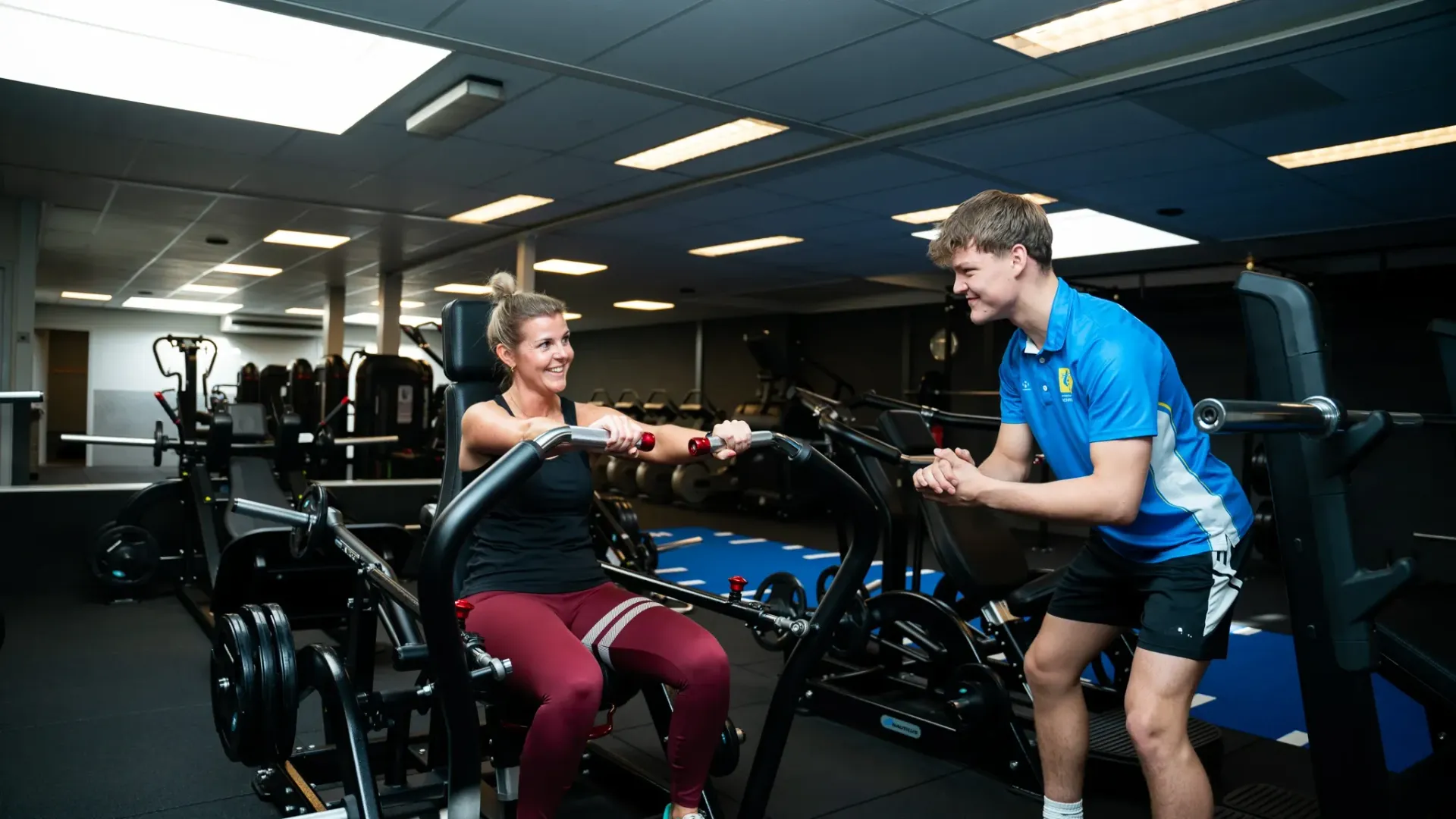 Een man helpt een vrouw met oefeningen op een apparaat in een sportschool.