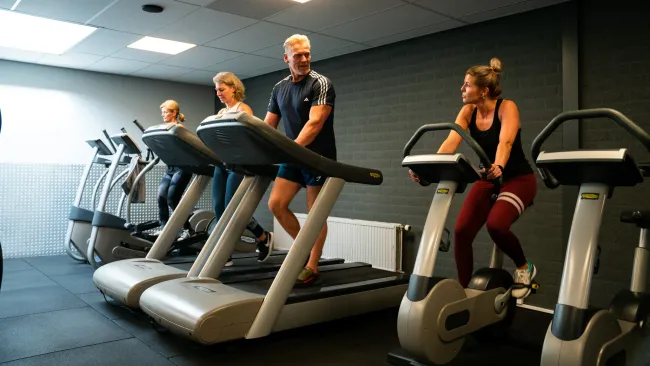 Een groep mensen rent op loopbanden, daarlangs fiets een vrouw op een hometrainer, daarlangs gebruikt een vrouw een elliptical trainer in een sportschool.