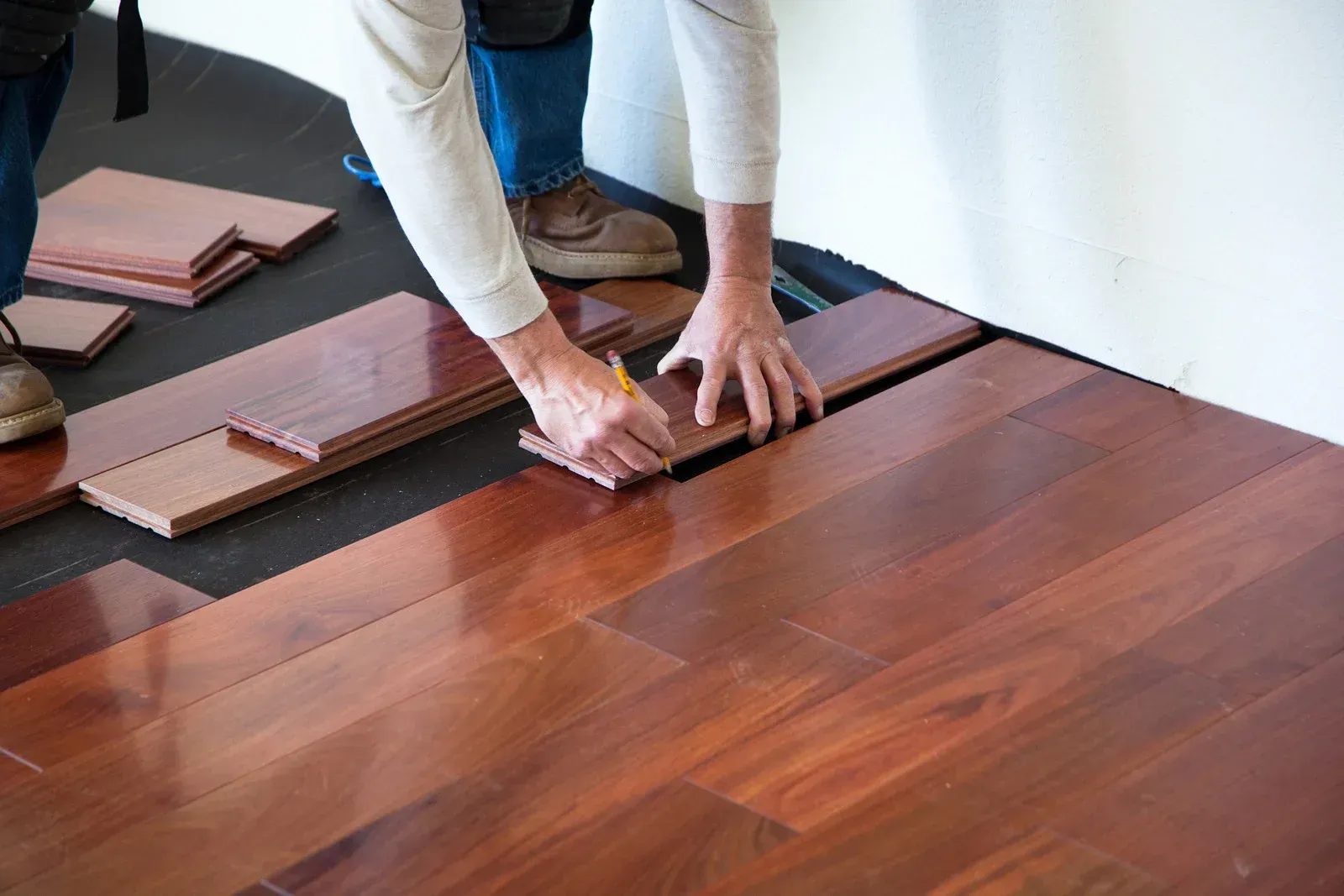 Person installing hardwood floor planks, using a pencil to mark a board for cutting.