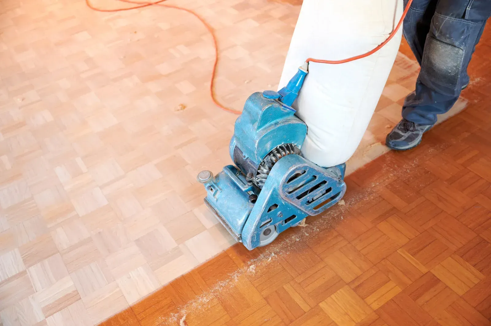 Person sanding a parquet wood floor with a blue machine; sawdust accumulates.