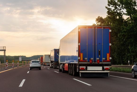 A row of semi trucks are driving down a highway at sunset.