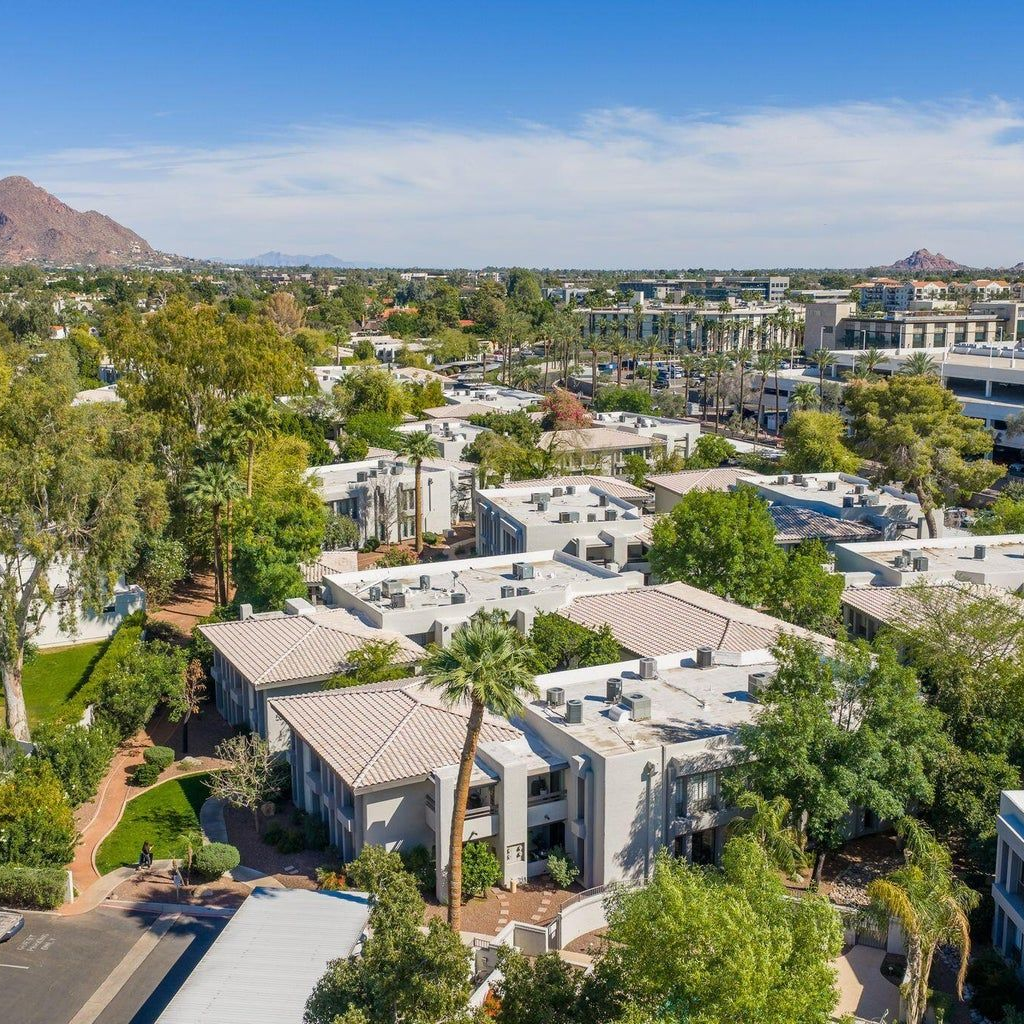 An aerial view of a multi-unit housing complex with light-colored buildings and tile roofs, surrounded by lush trees.