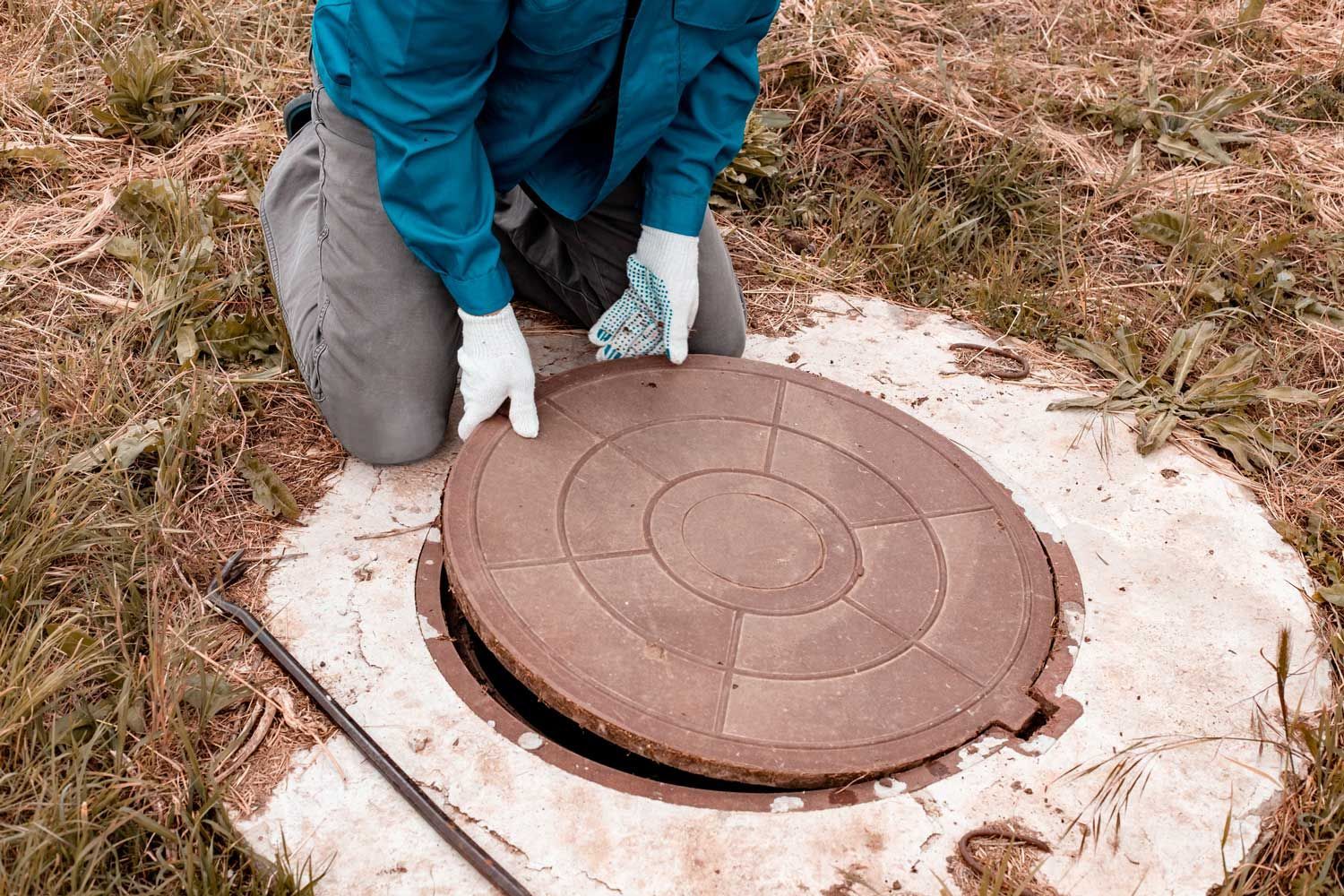 Person opening a septic tank cover in a grassy area, wearing gloves and blue shirt.