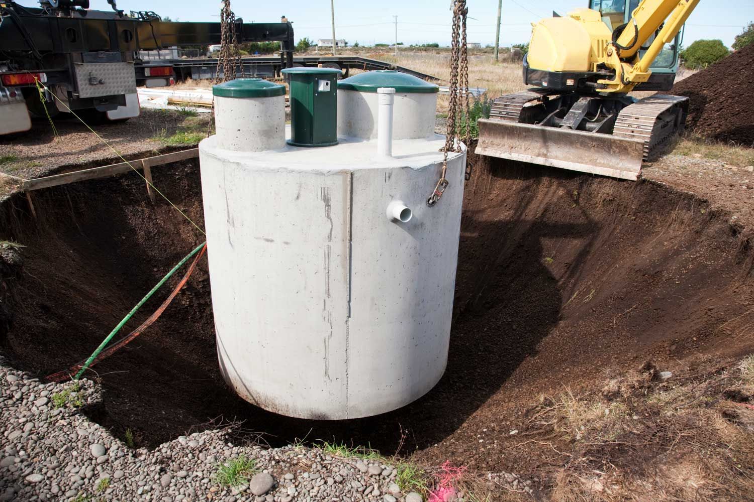 Concrete septic tank being lowered into a large excavated hole by a crane; construction site.