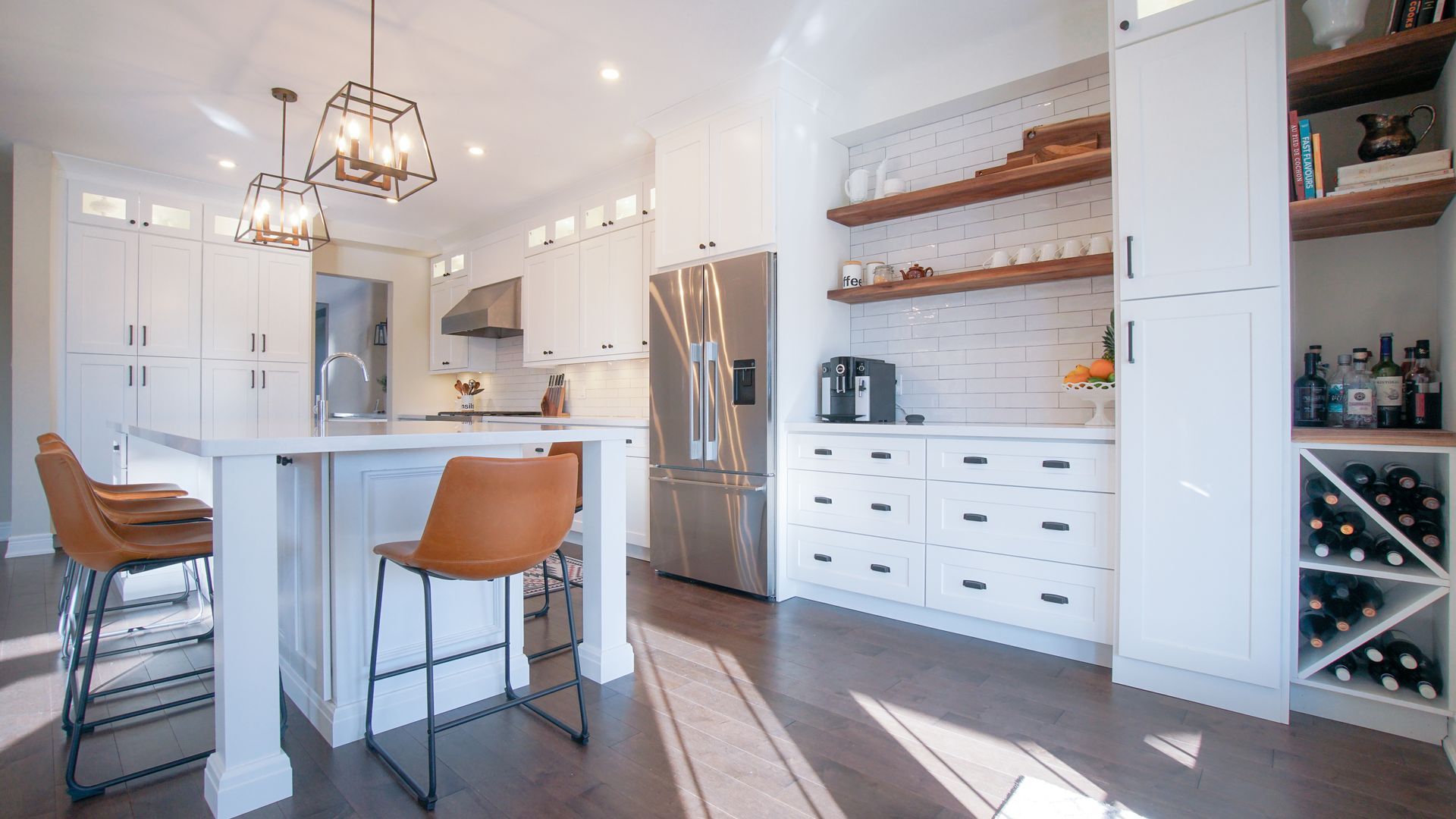 Bright, modern kitchen with white cabinets, island, and stainless steel appliances. Brown stools and wood shelves add warmth.