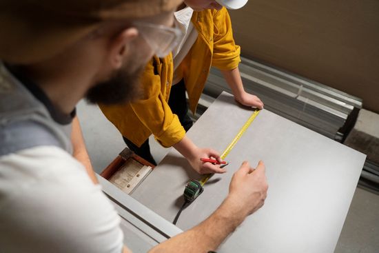 A man and a woman are measuring a piece of wood with a tape measure.