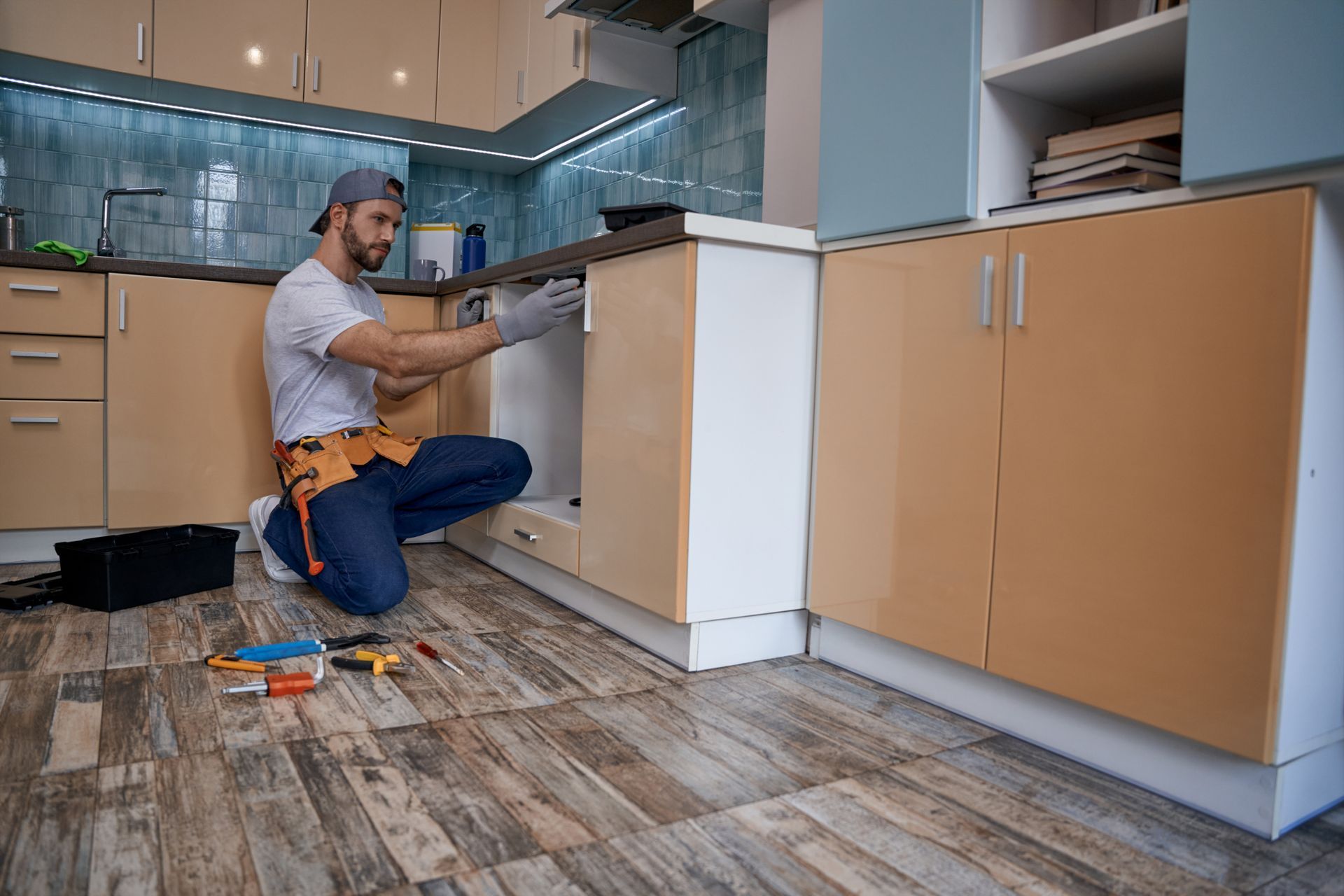 A man is kneeling on the floor in a kitchen fixing a sink.