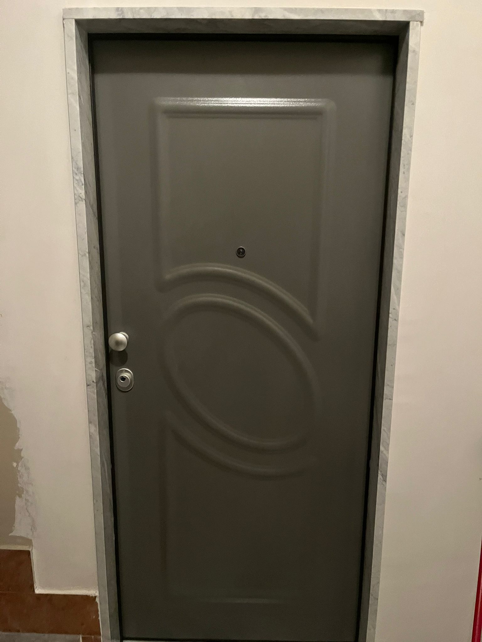 Porta in metallo grigio con design ovale e ferramenta argentata, inserita in un telaio grigio e parete bianca.