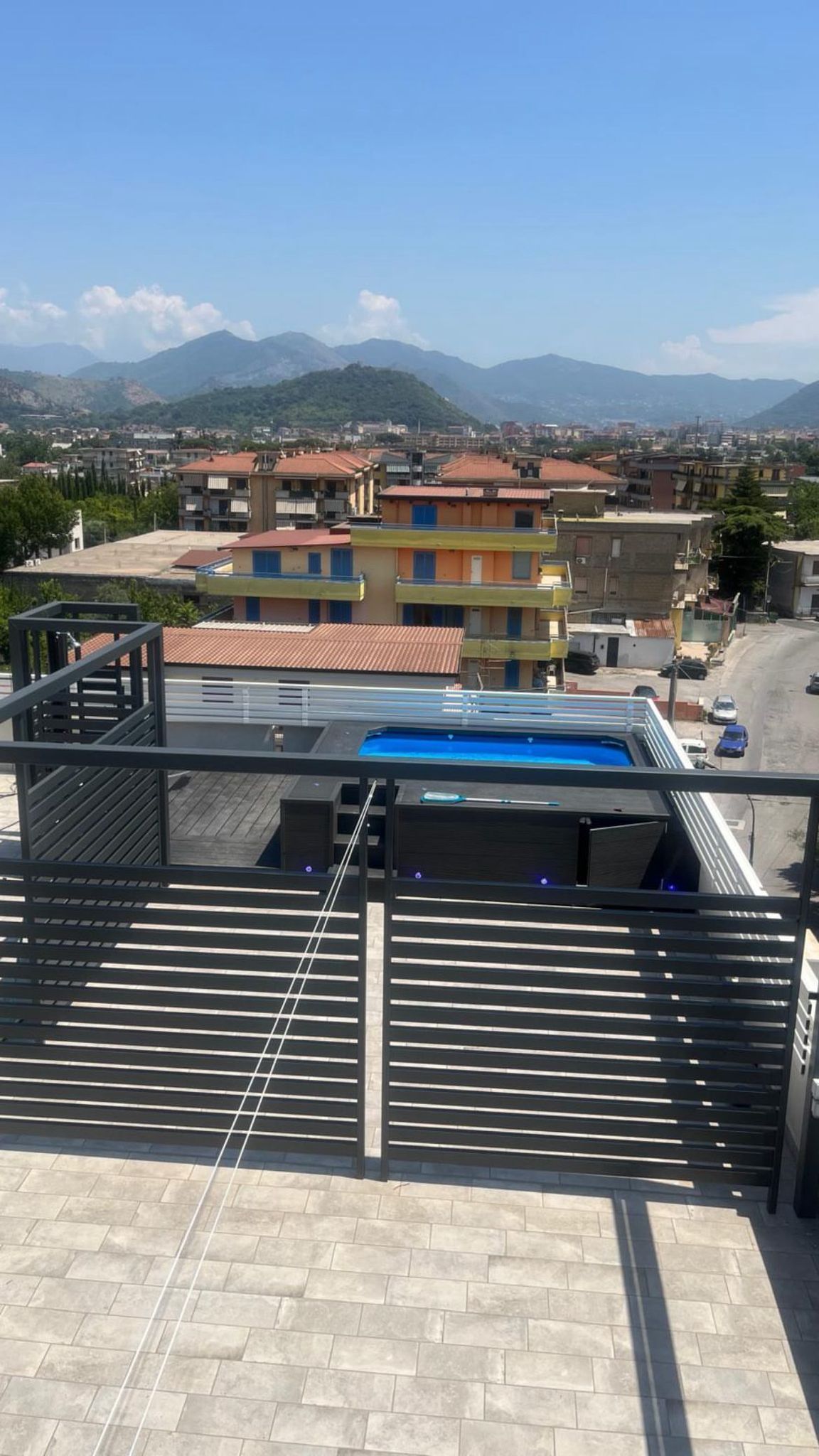 Vista di un paesaggio urbano con montagne sullo sfondo, una piscina e un cancello metallico.