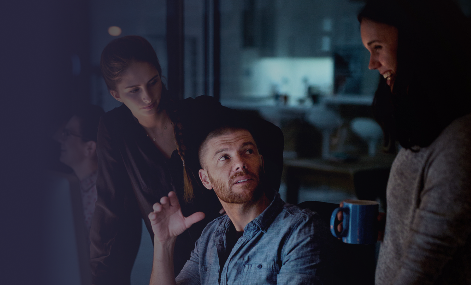 Three people in a dimly lit room. A man gestures, looking upward. Two women listen and smile.