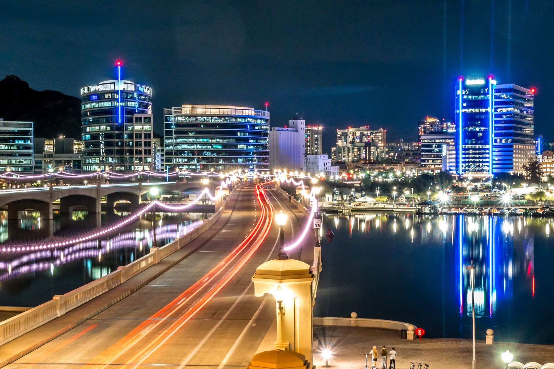 Nighttime city skyline with illuminated buildings, bridge traffic light trails, and reflections on the water
