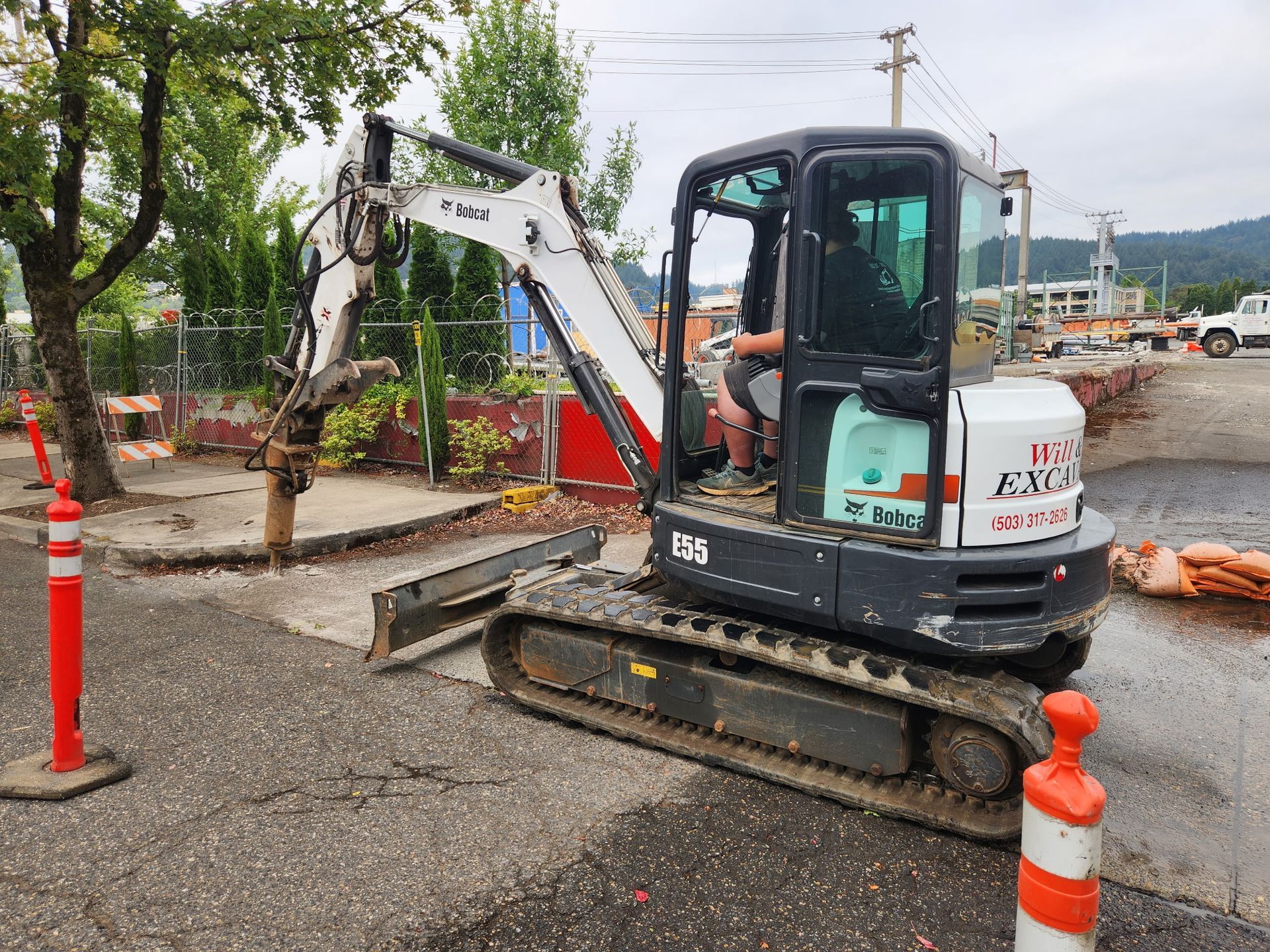 Mini excavator on asphalt. Operator in cab. Construction site with red/white traffic posts, trees.
