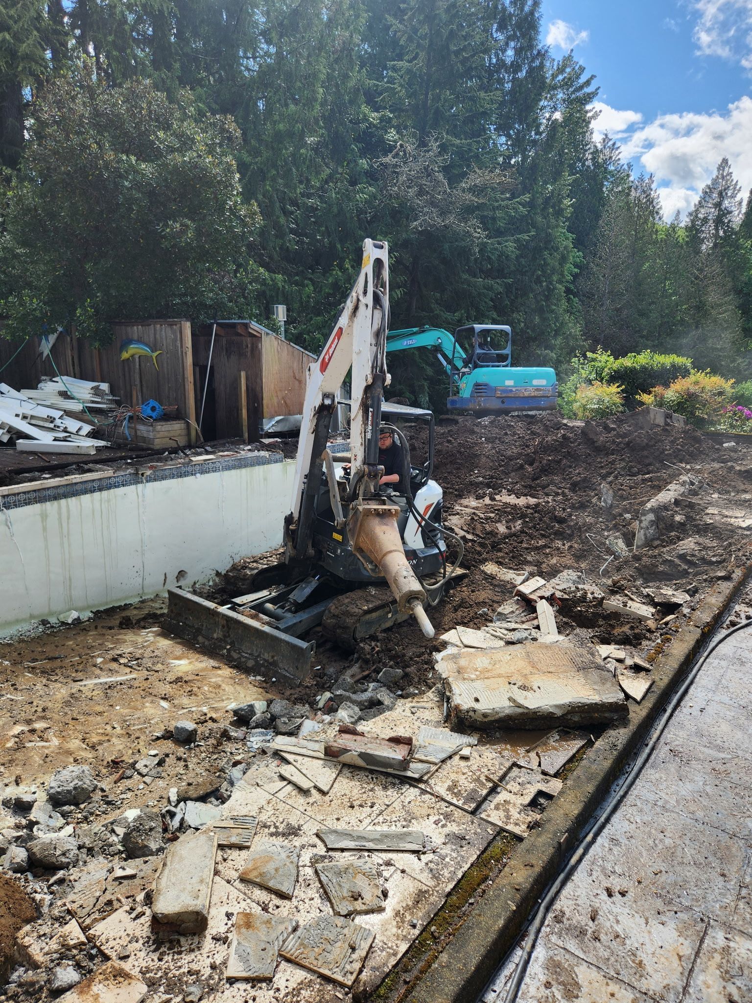 Mini excavator demolishing concrete near a retaining wall, with a second excavator in the background.